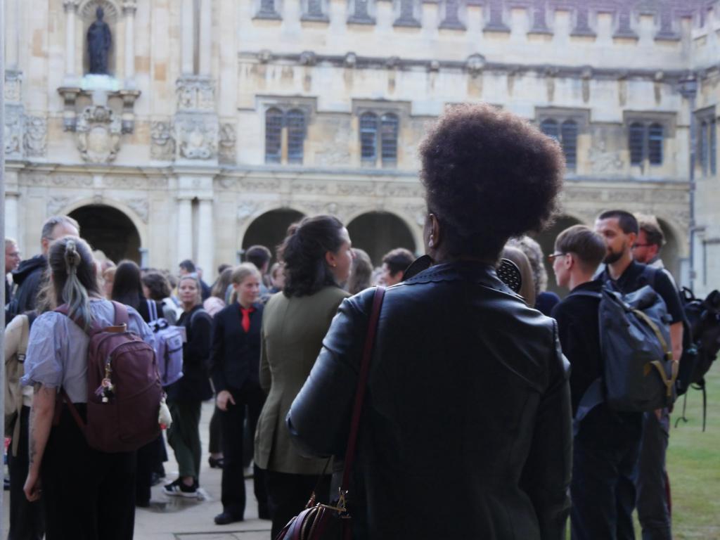 view of the backs of a crowd of (predominantly white) people in an Oxford quad, with a black person in the foreground, also seen from the back