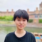 Photo of an East Asian person with short black hair wearing a black t-shirt with Magdalene College and the River Cam in the background