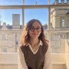 White woman in a shirt, sweater vest and glasses sitting in front of a window, from which the walls of an Oxford college can be seen.